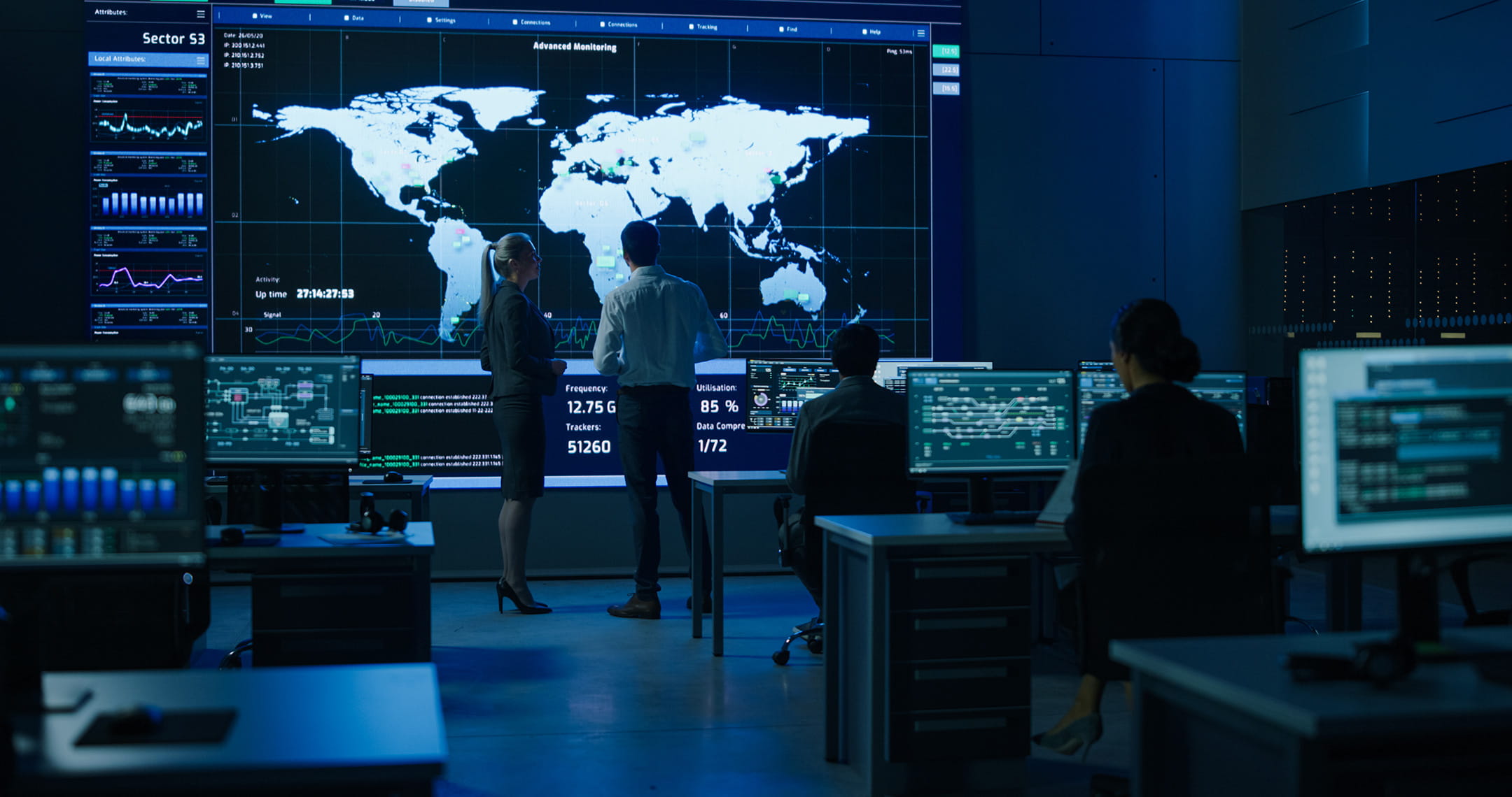 Operations control room with large wall display showing a global map, live telemetry, and network activity graphs; analysts at computer workstations monitor data while two staff review the central screen.