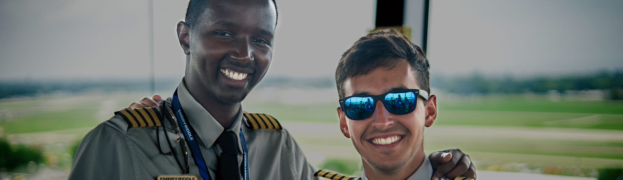 Two pilots stand together in the airport in front of a window overlooking the flight field.