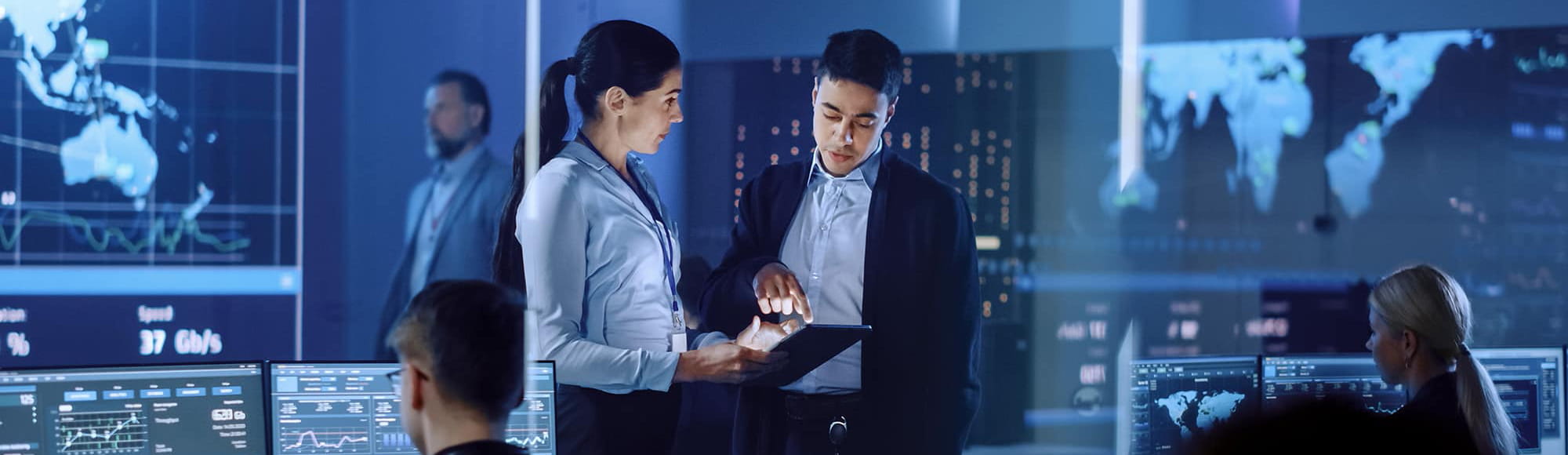 two people look at a tablet together in a control room with maps showing on large screens behind them