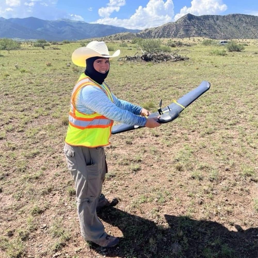John Christensen stands in a field surrounded by mountains while holding an uncrewed system and wearing a safety vest and cowboy hat