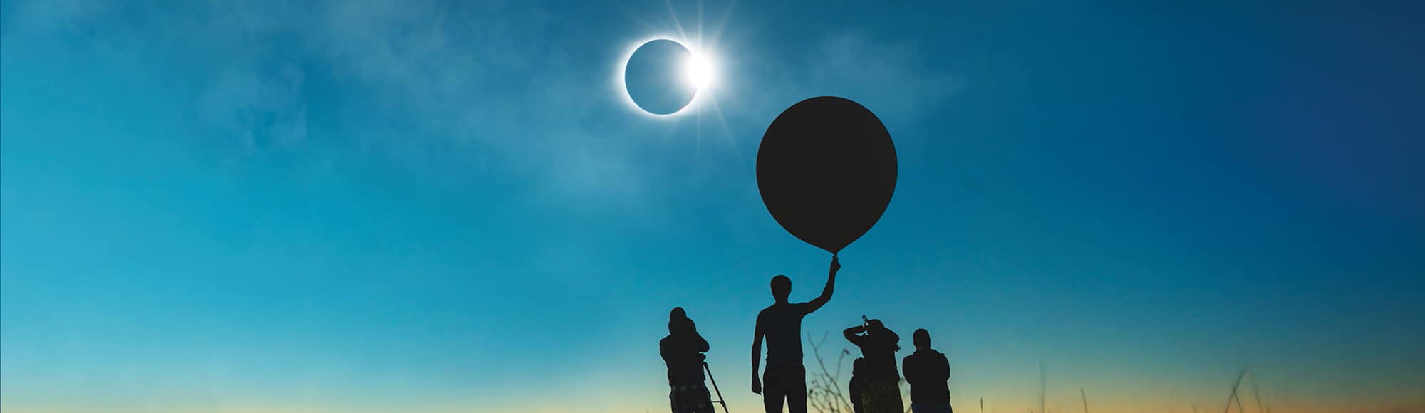 A group of people stand in a field observing the solar eclipse while holding a weather balloon