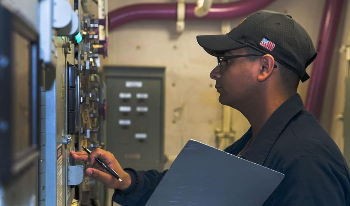 A person carrying a clipboard looks at a wall of switches and controls.