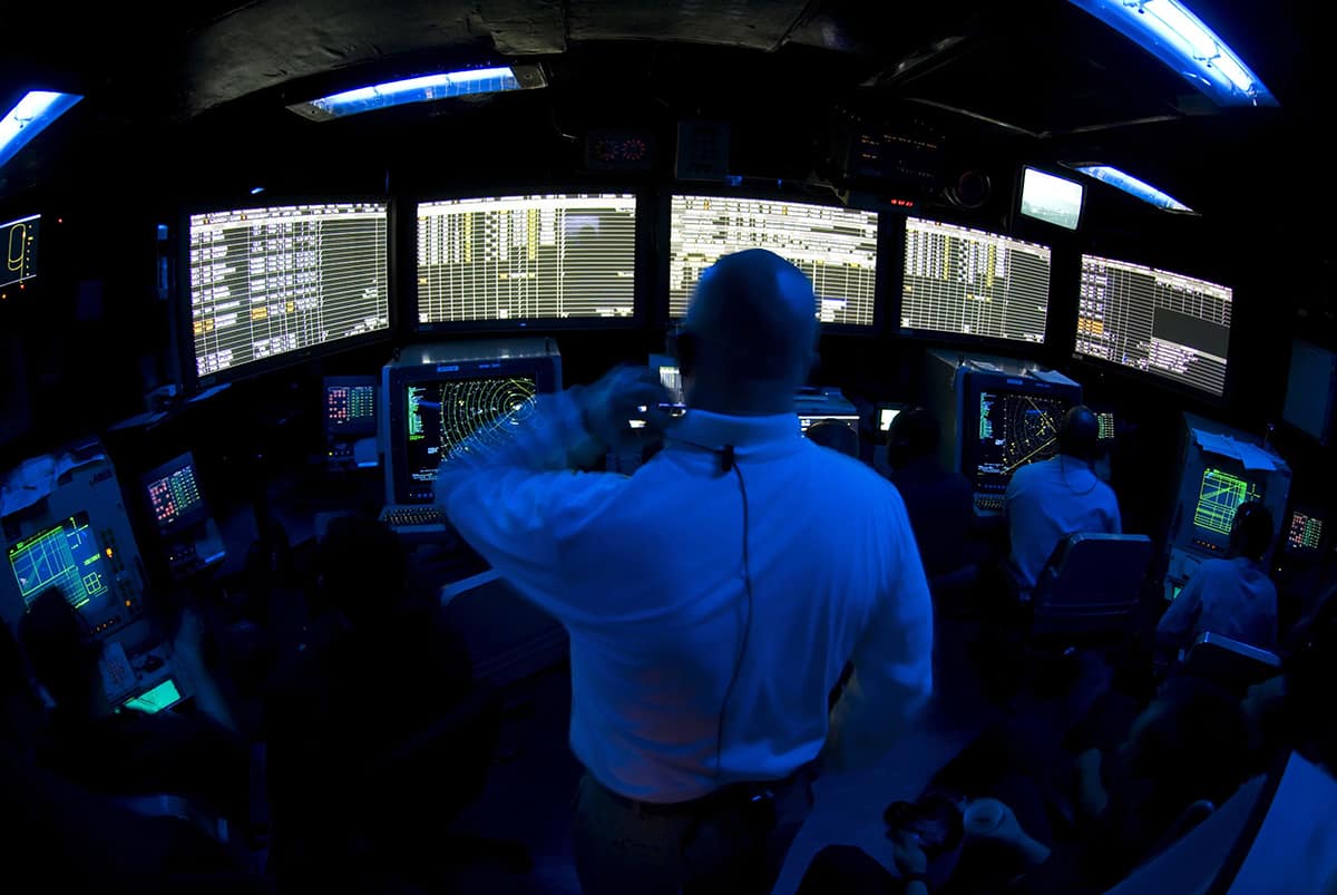 Man in white shirt stands in room with blue lighting looking at five large computer screens.