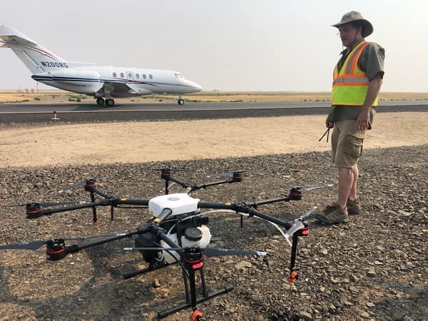 Man stands next to drone next to runway with airplane in background.
