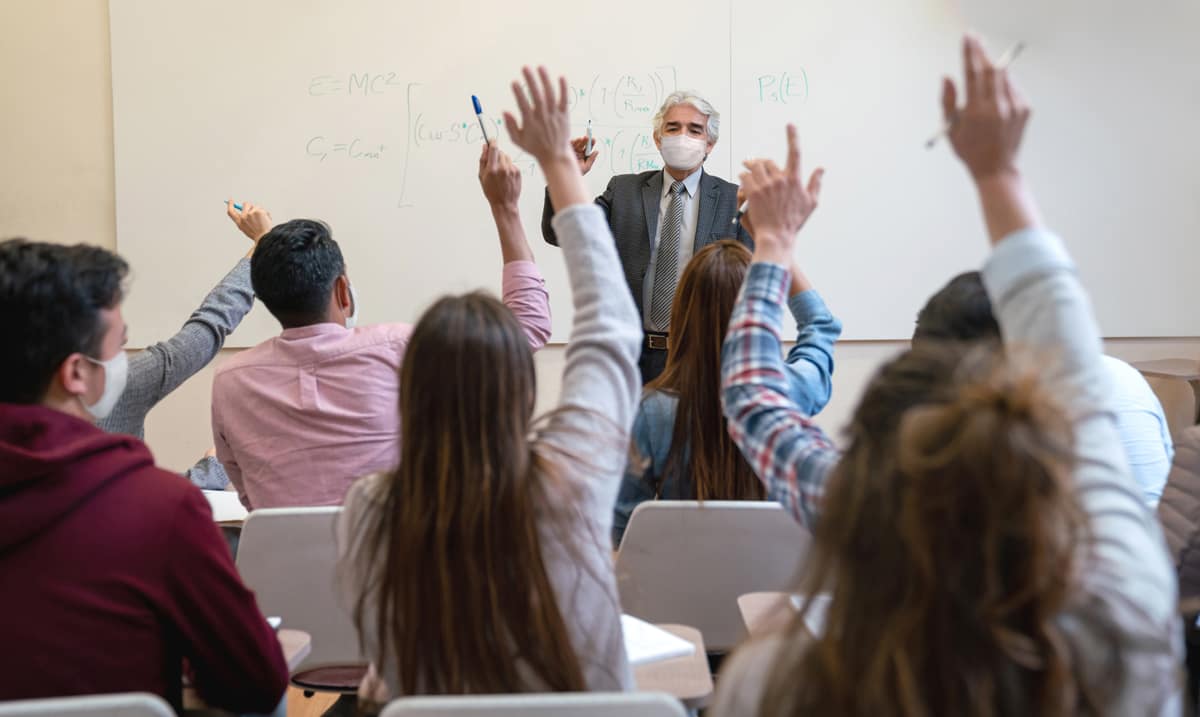 Students raise hands in classroom while professor wears medical mask.