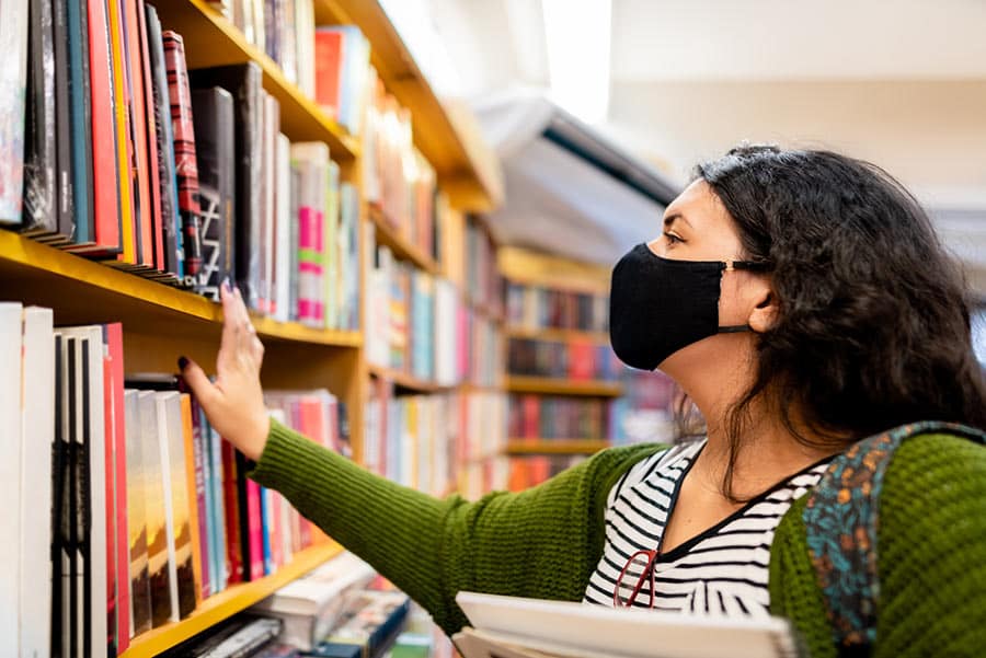 Student wearing medical mask looks at books in library.