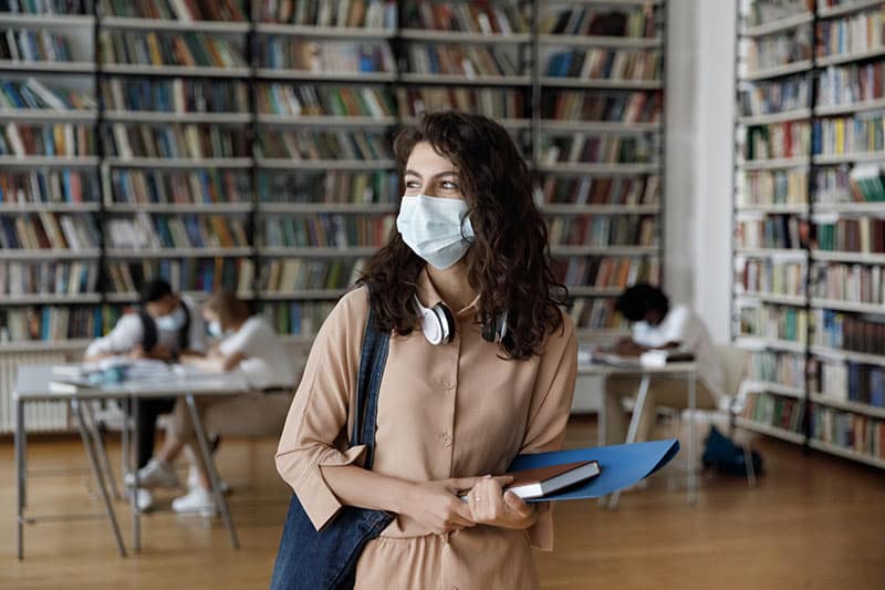 Women stands in library holding book and wearing surgical mask.