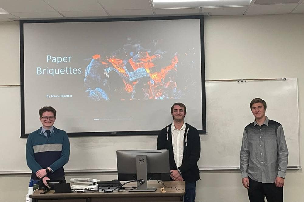 Three students stand in front of white board and projector screen.