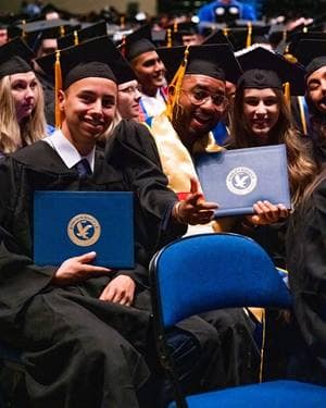 Students in cap and gown hold up diplomas at graduation ceremony.