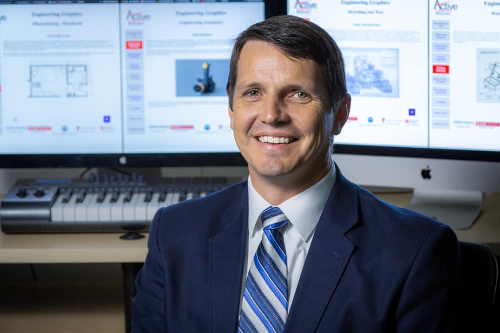 Dr. Jeremy V. Ernst sits in front of computer monitors while wearing suit.