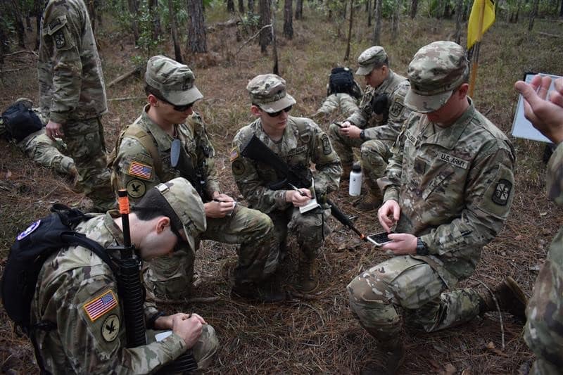 Army ROTC students write on notepads in woods.