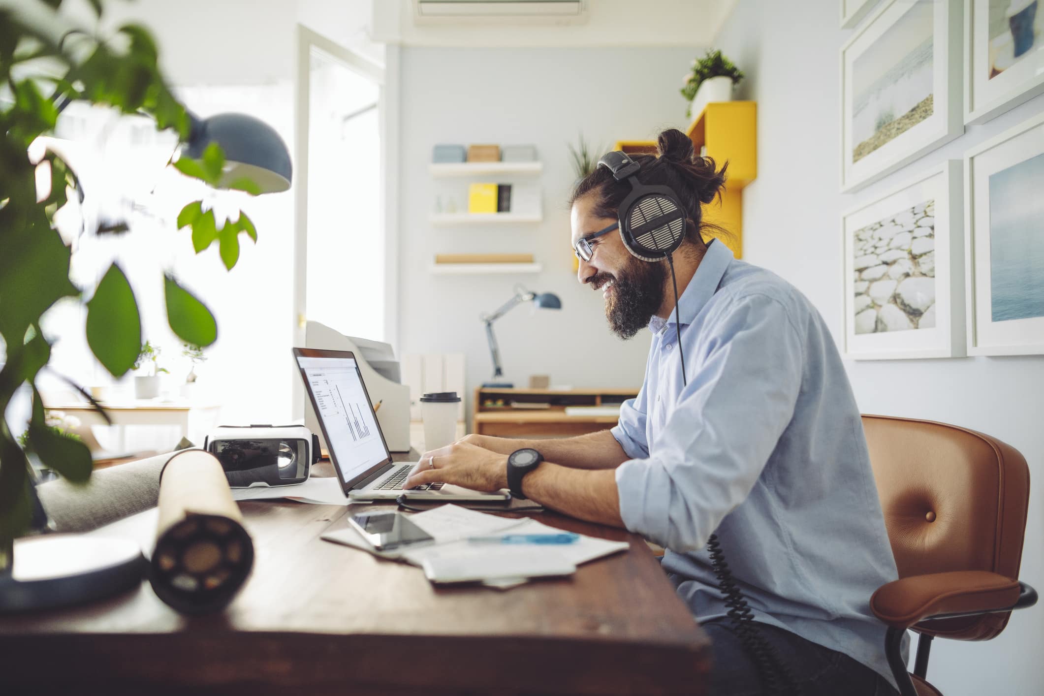 Man wearing headphones sits at desk and works on laptop in his home.