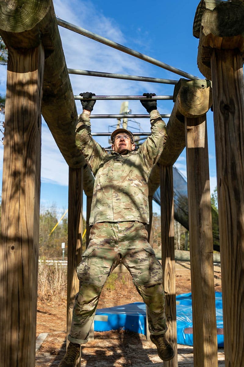 Army ROTC cadet hangs from bar on obstacle course.