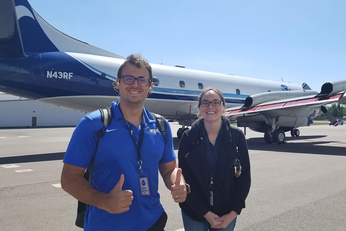 Man and woman pose on runway in front of airplane.