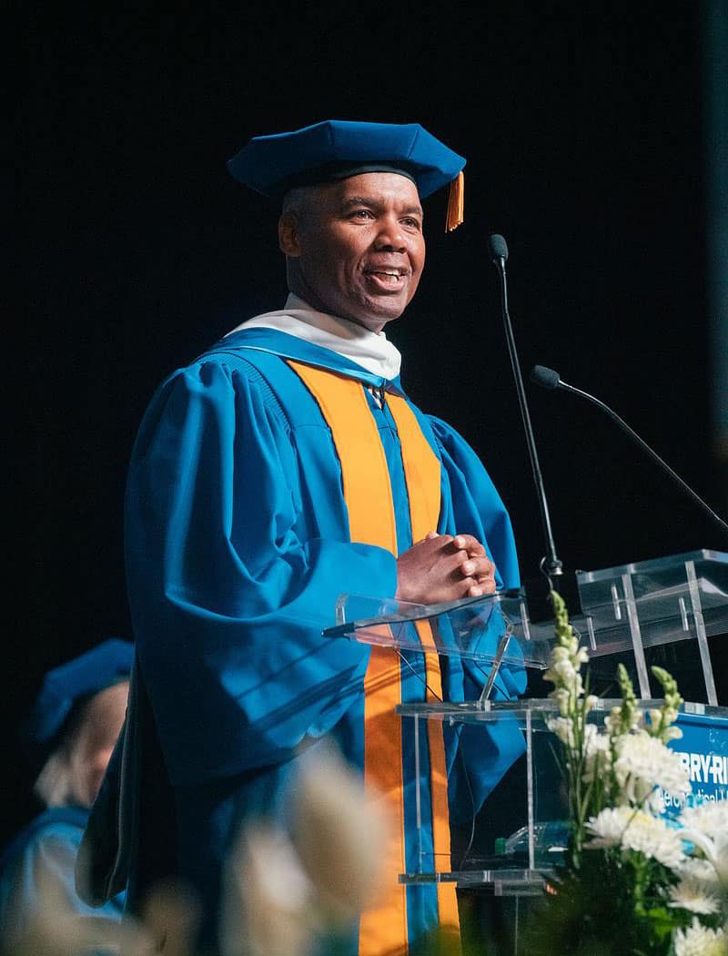 Man in cap and gown speaks at graduation ceremony.