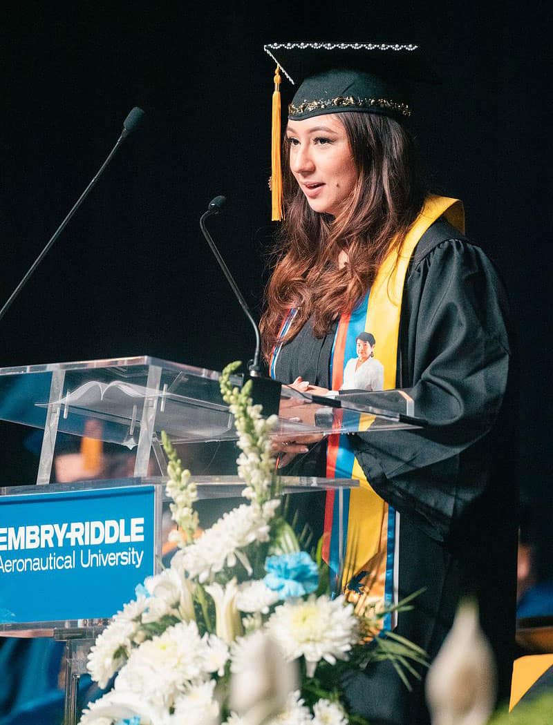 Woman in cap and gown speaks at graduation ceremony.