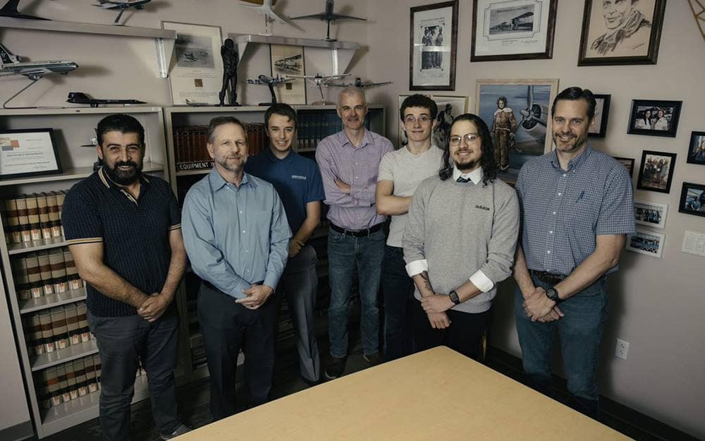 Seven men pose behind a table in a office with multiple model plans are on shelves and pictures hanging on the walls.