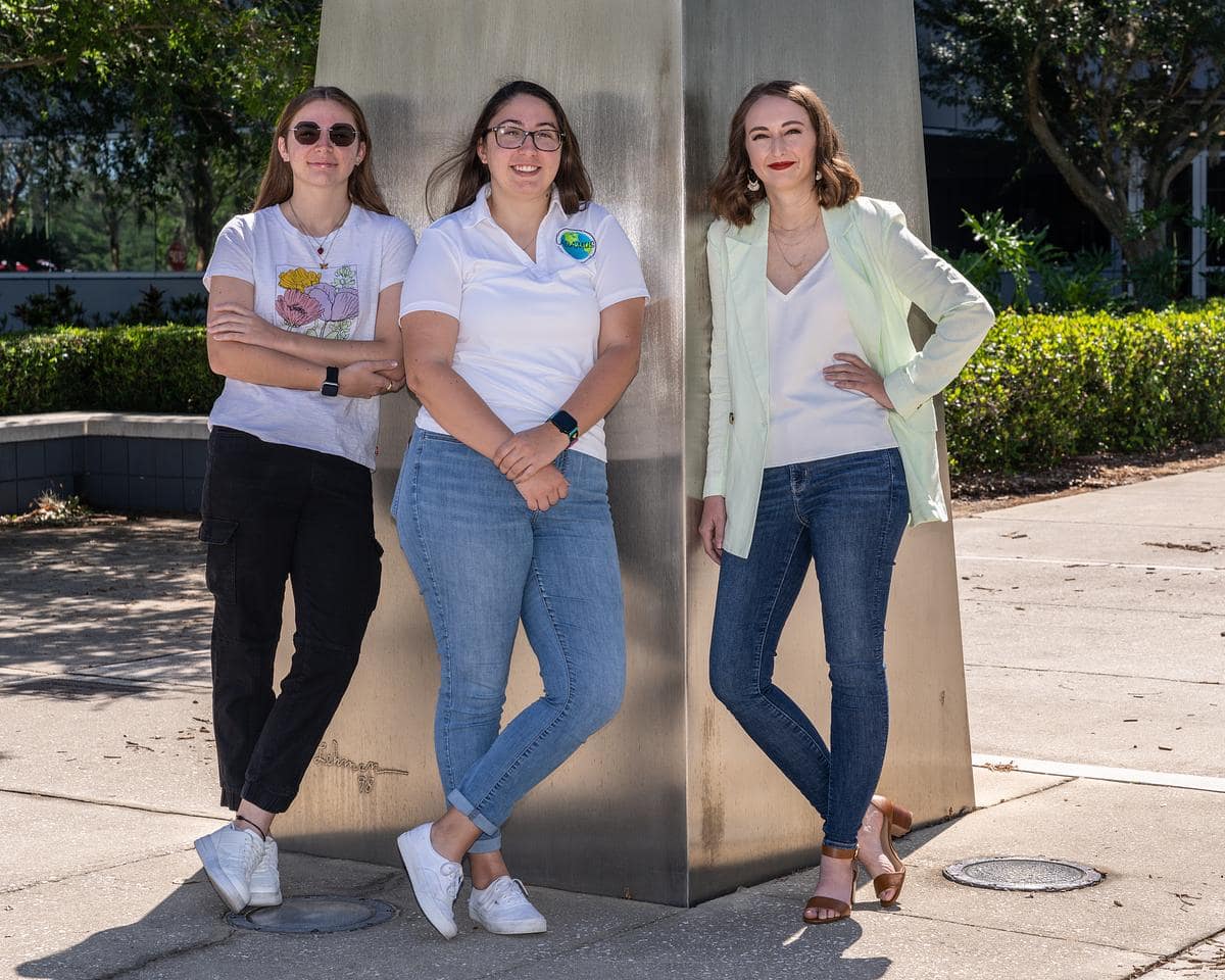 Three women pose for picture next to metal pillar.