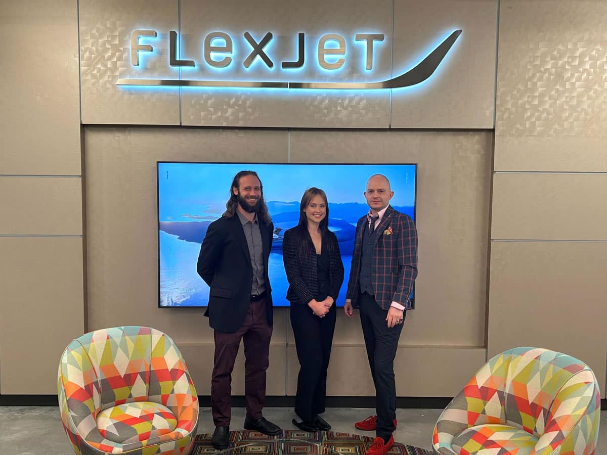 Three people pose in front of TV screen below a Flexjet logo.