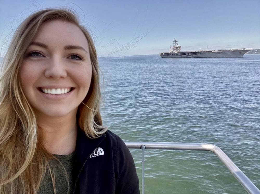 Woman poses for picture in front of warship in harbor.