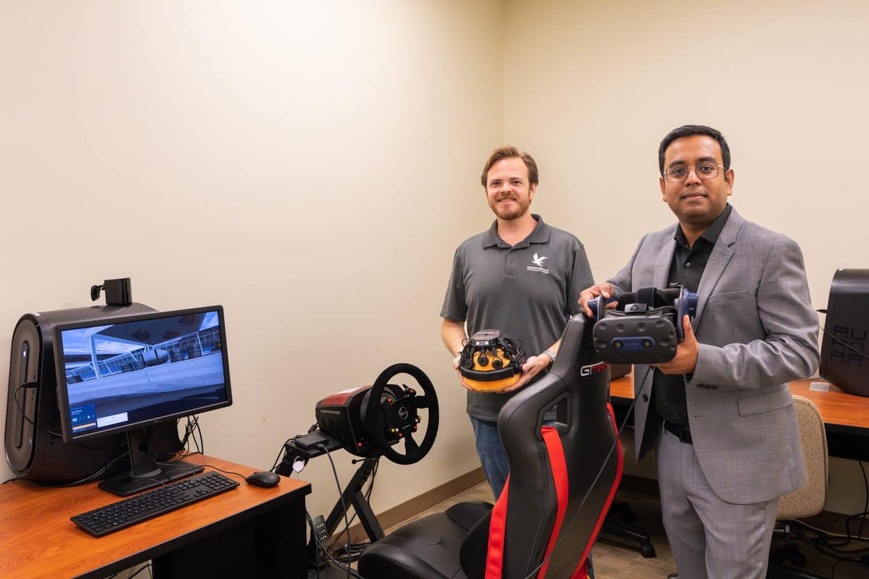 Two men hold VR driving simulator next to computer.