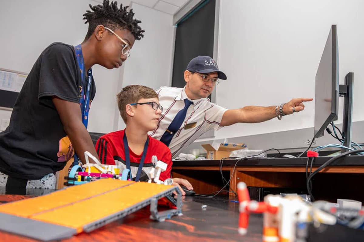 Man points at computer monitor while two students look on.