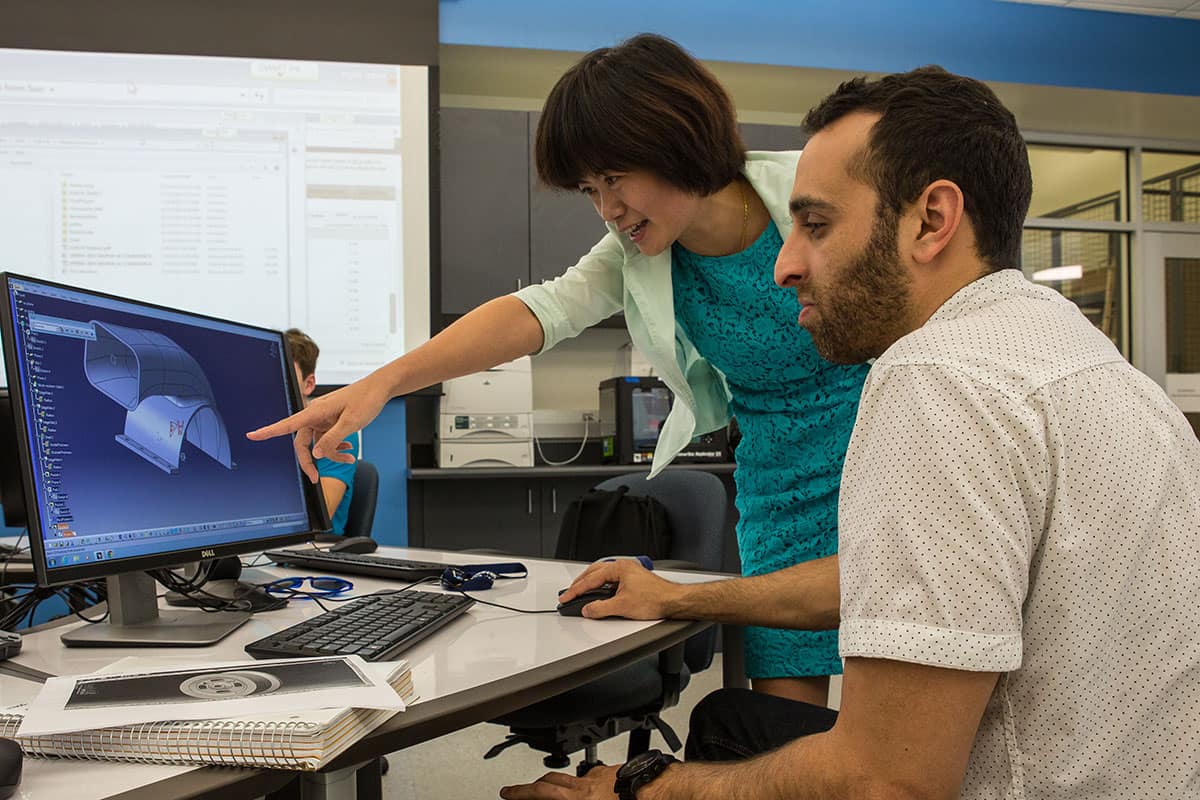 Woman points at computer monitor while man looks on sitting at desk.