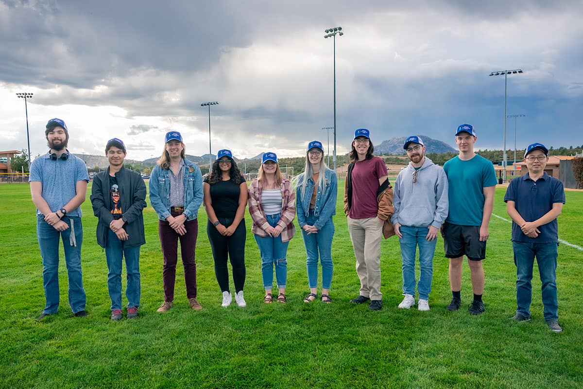 10 students stand on a green athletic field with desert mountains and stormy skies in the background.