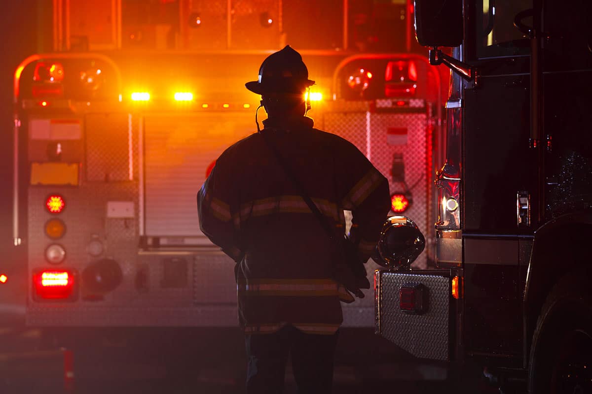 A firefighter infront of a truck flashing lights at night.