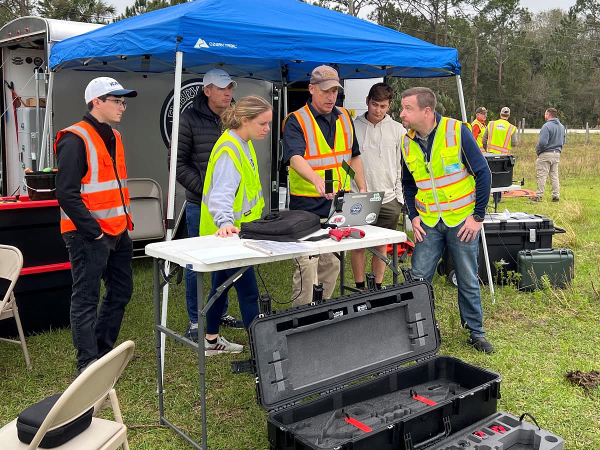 Six people stand under tent looking at laptop.