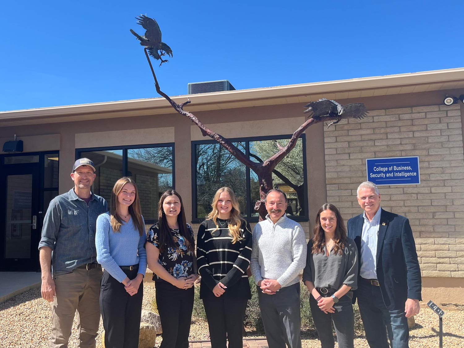 Seven students and faculty standing in front of a sculpture of a tree with two eagles; signage reads College of Business, Security and Intelligence on the building behind them.