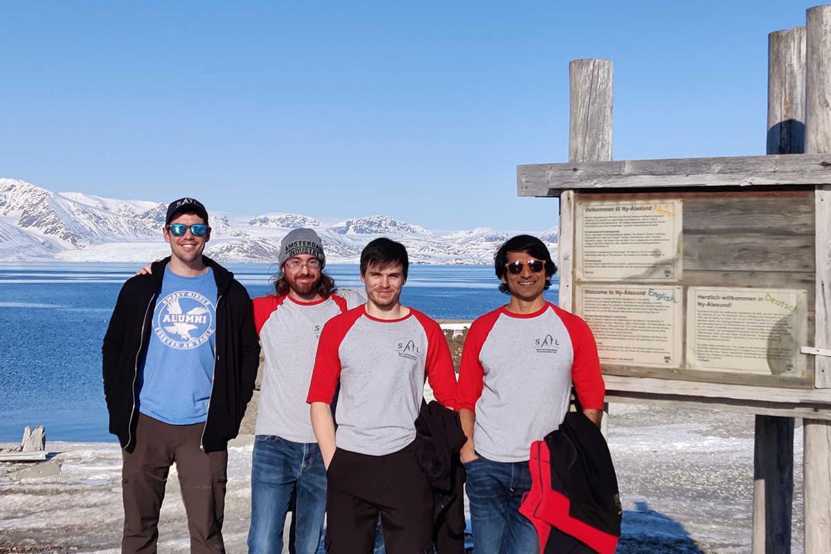 Four students posing outdoors near coastal research site with snow-covered mountains and ocean in background.