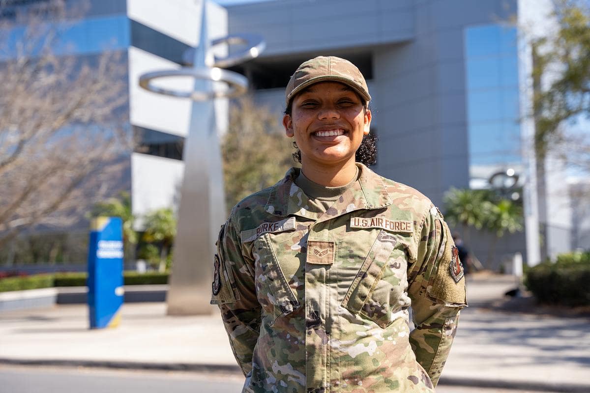 U.S. Air Force airman in camouflage uniform stands in front of a modern campus building with a metal ring sculpture, name tape 'Burke' visible.