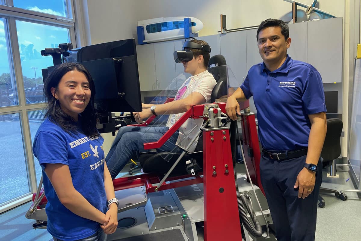 Embry‑Riddle researcher Dr. Hever Moncayo (right) and graduate student Gabriela Gavilánez Gallardo (left) stand in the Advanced Dynamics and Control Lab next to the flight simulator being used for their AI research project. Aerospace engineering Ph.D. student Nathan Schaff, who is also a pilot, is sitting in the flight simulator. (Photo: Embry‑Riddle/Melanie Azam)