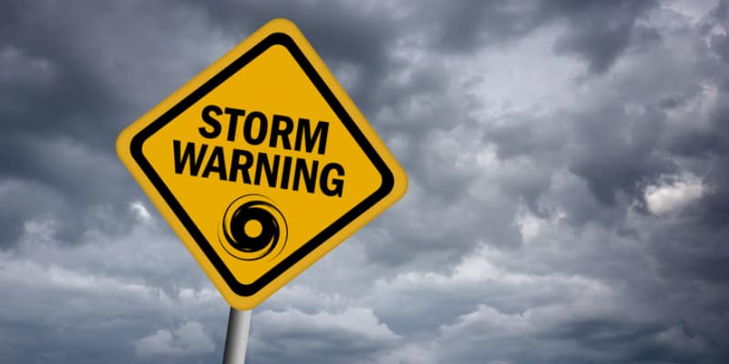Yellow diamond road sign reading Storm Warning with hurricane symbol, set against dark storm clouds.
