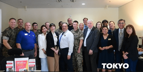 Group of 20 students, faculty, and military personnel posing together indoors with table display, with 'TOKYO' text in corner.