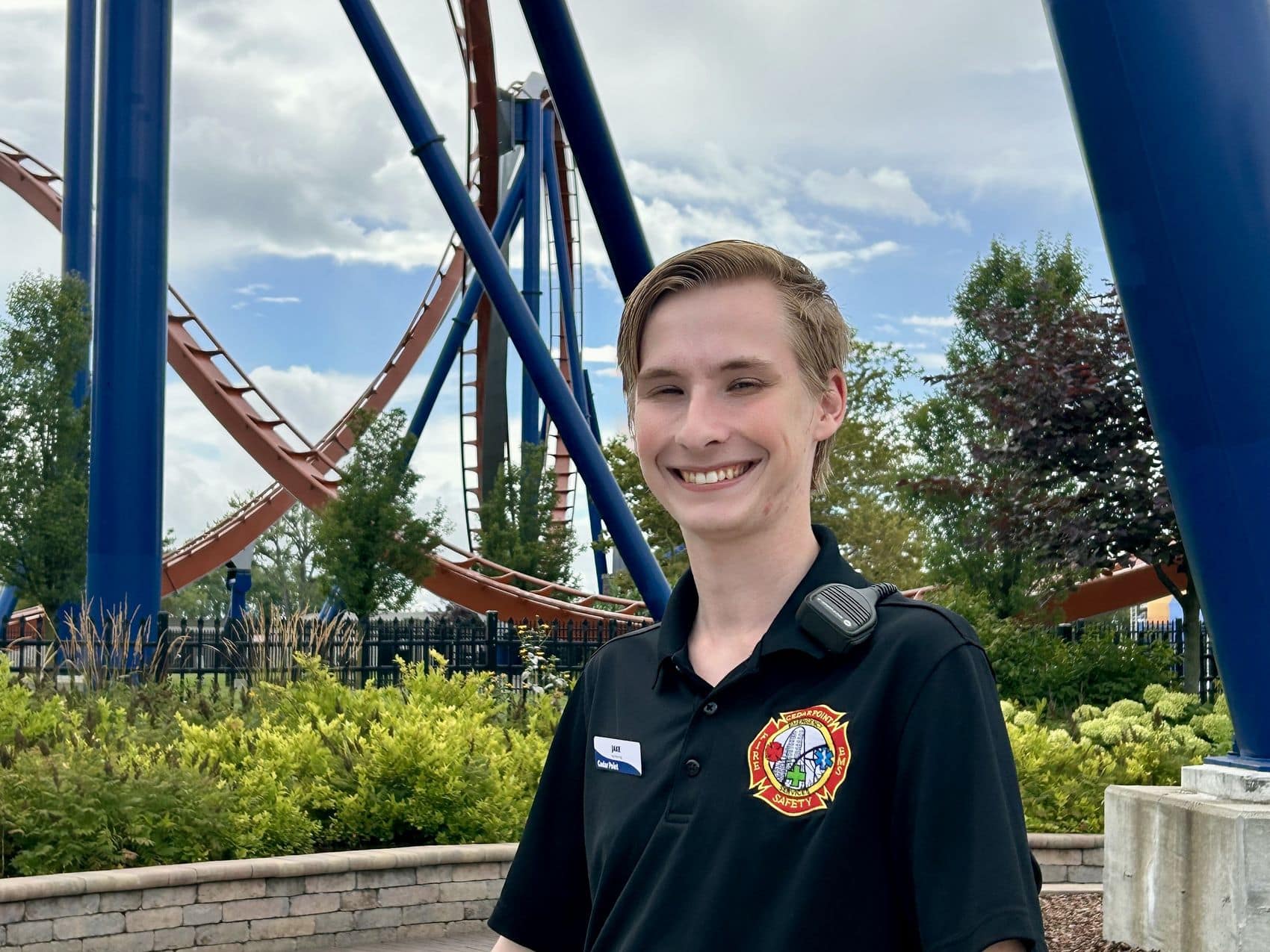 Jacob Welch in a Cedar Point fire, EMS, safety uniform standing in front of roller coaster with looping track and support structures in amusement park.