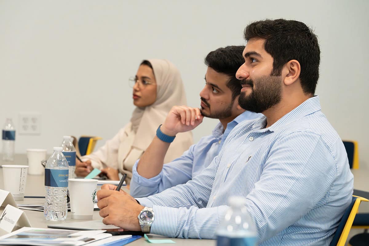 Students seated at classroom table taking notes and listening attentively.