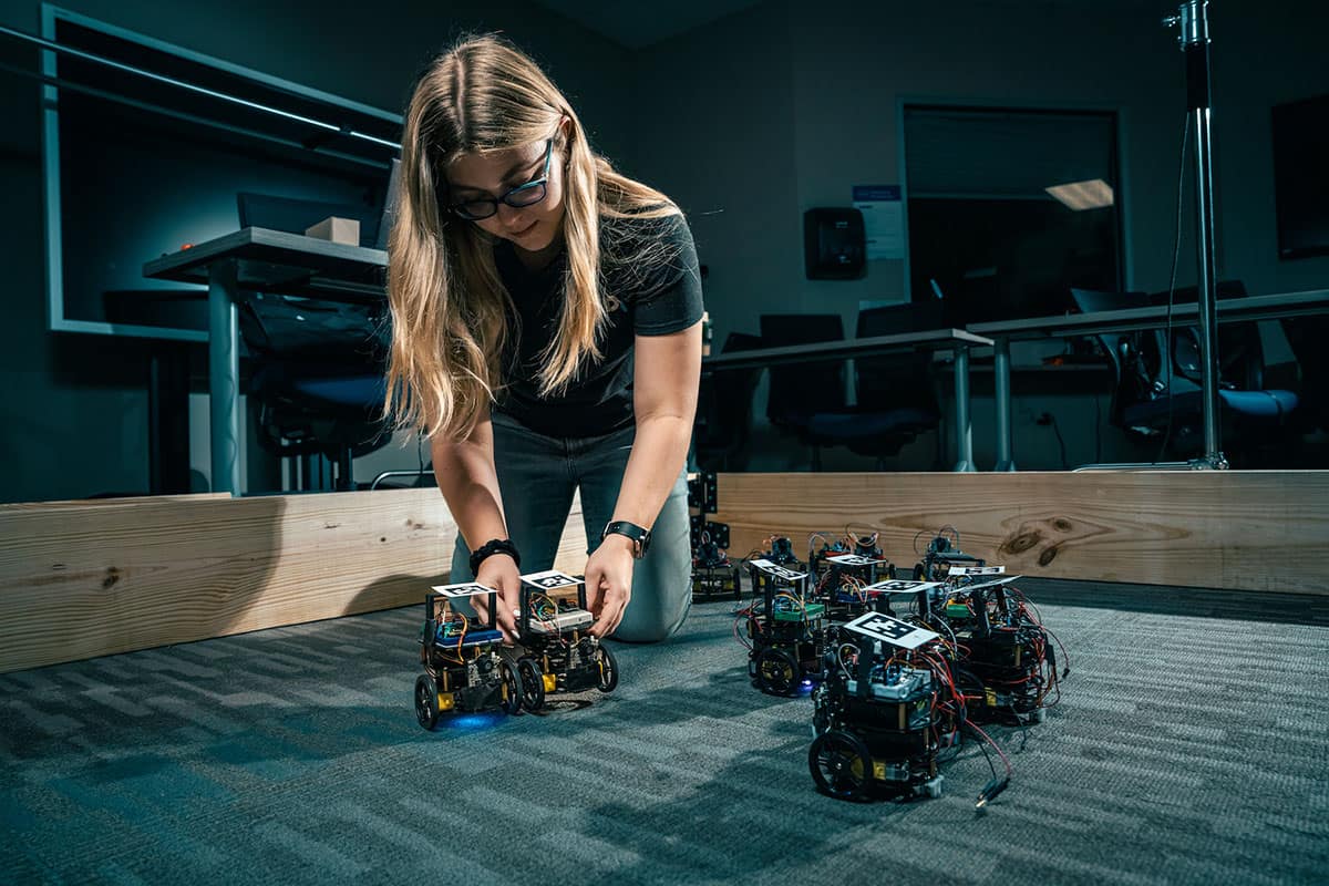Student researcher Noa Teed, a senior majoring in Software Engineering, adjusts robotic vehicles programmed to interact at timed intervals and change each other's internal codes. (Photo: Embry‑Riddle/William Fredette-Huffman)