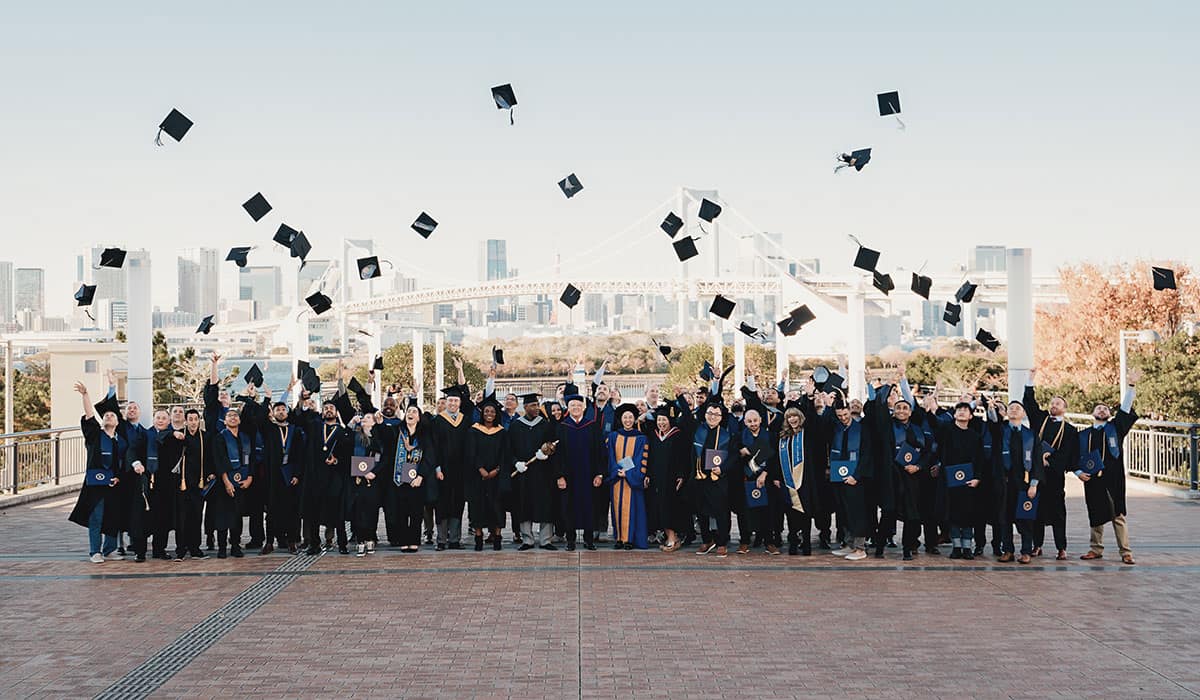 Two dozen graduates in caps and gowns celebrating and tossing caps in the air with waterfront city skyline and suspension bridge in background.