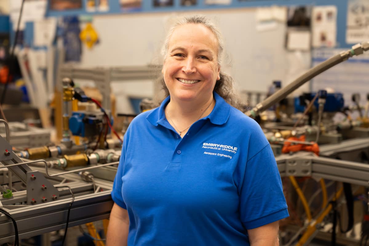 Dr. Girard in an Embry-Riddle Aerospace Engineering polo poses in a lab with fluid systems test rig, piping, valves, and instrumentation equipment.