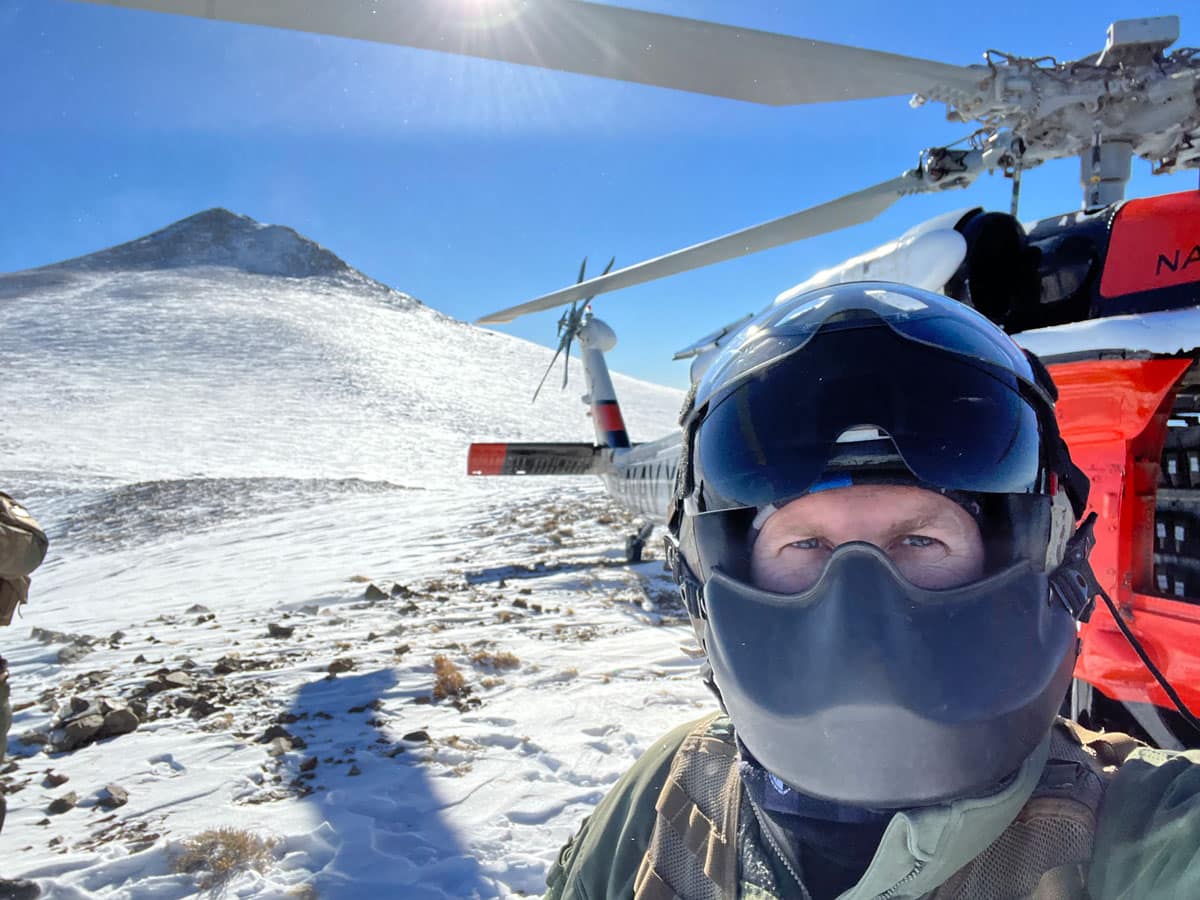 Person wearing a helmet and face covering taking a selfie beside a helicopter with rotor blades on a snowy mountain terrain.
