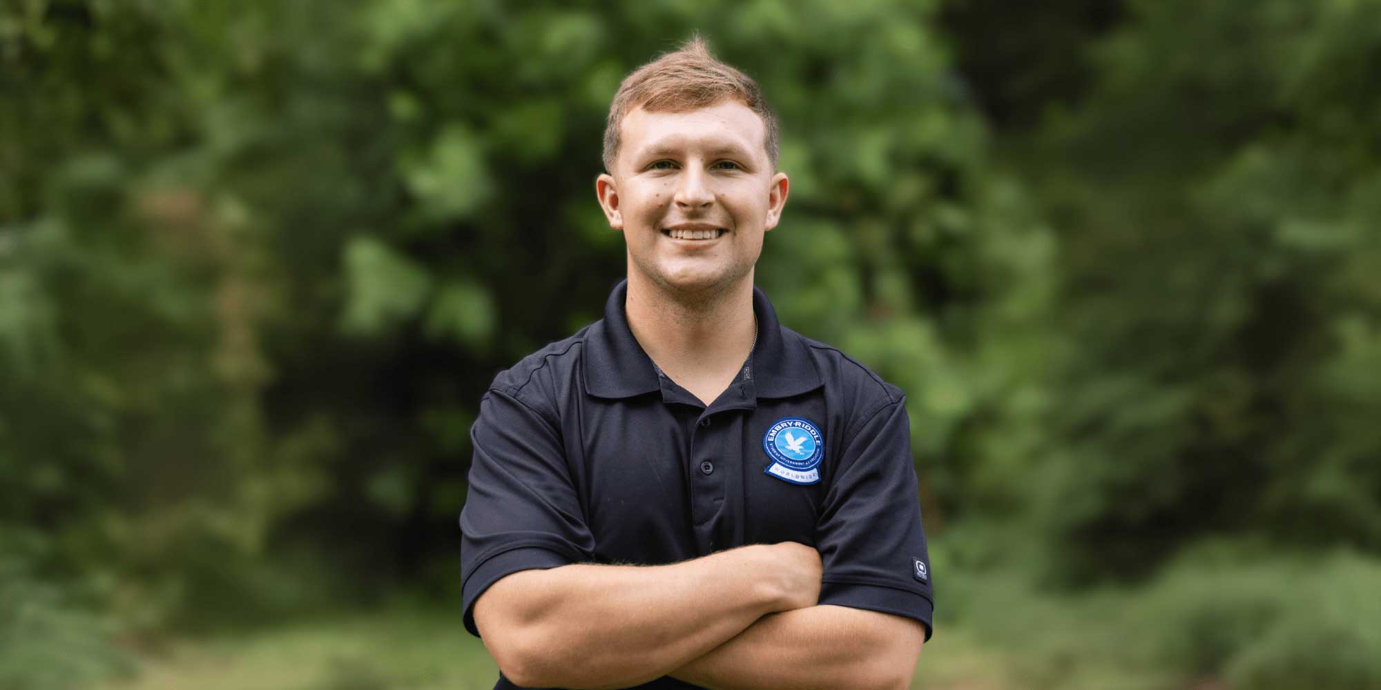 Evan stands outdoors in a dark polo embroidered with an Embry-Riddle seal, arms crossed, smiling in front of a leafy green background.