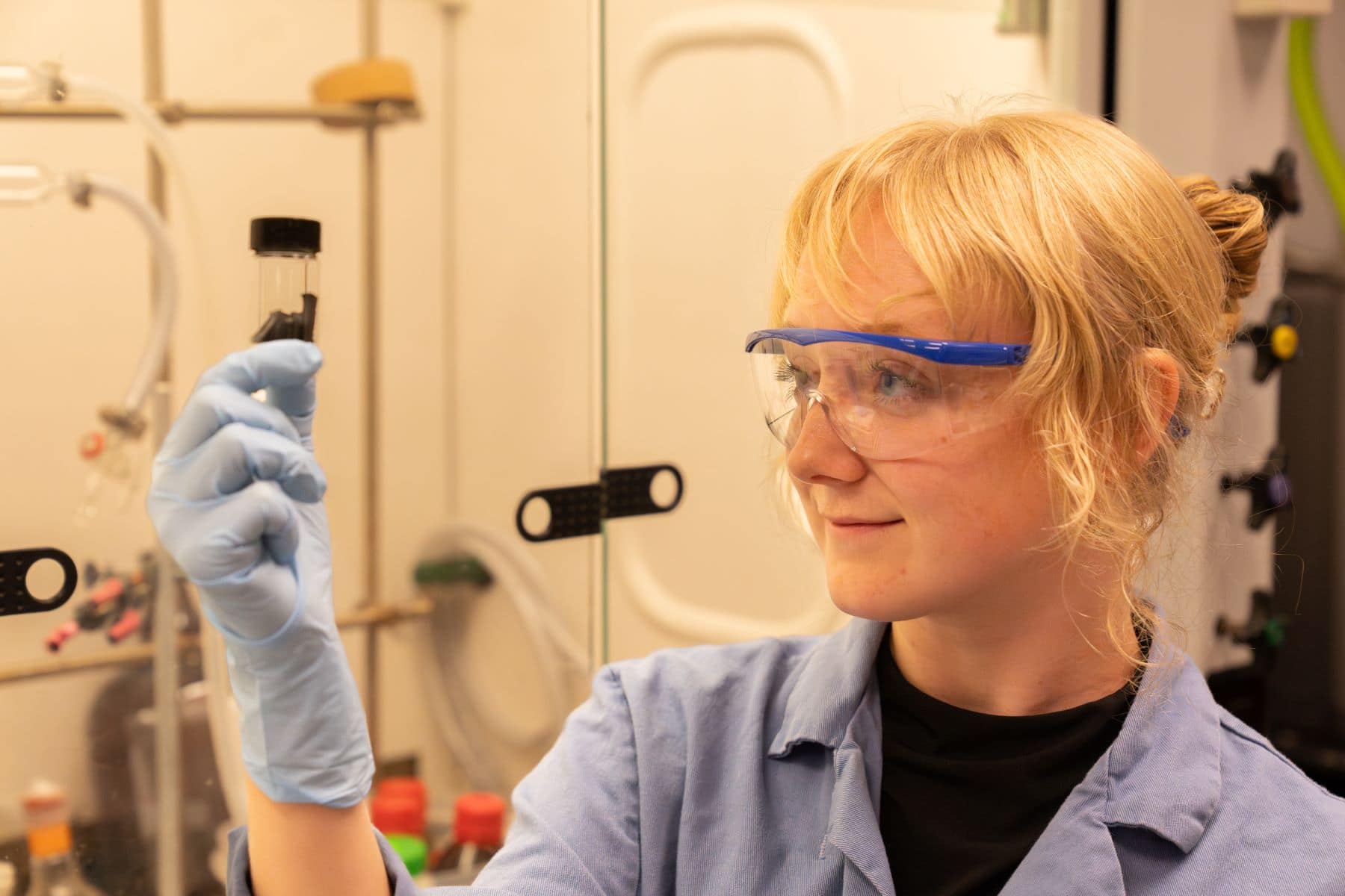 Scientist wearing safety goggles and gloves examines a small vial in a laboratory.