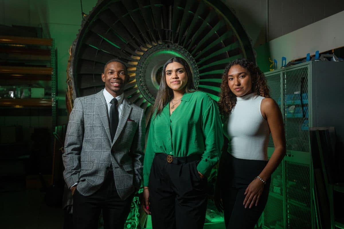 Three students in business wear pose in front of a large jet engine inside a lab or workshop space.