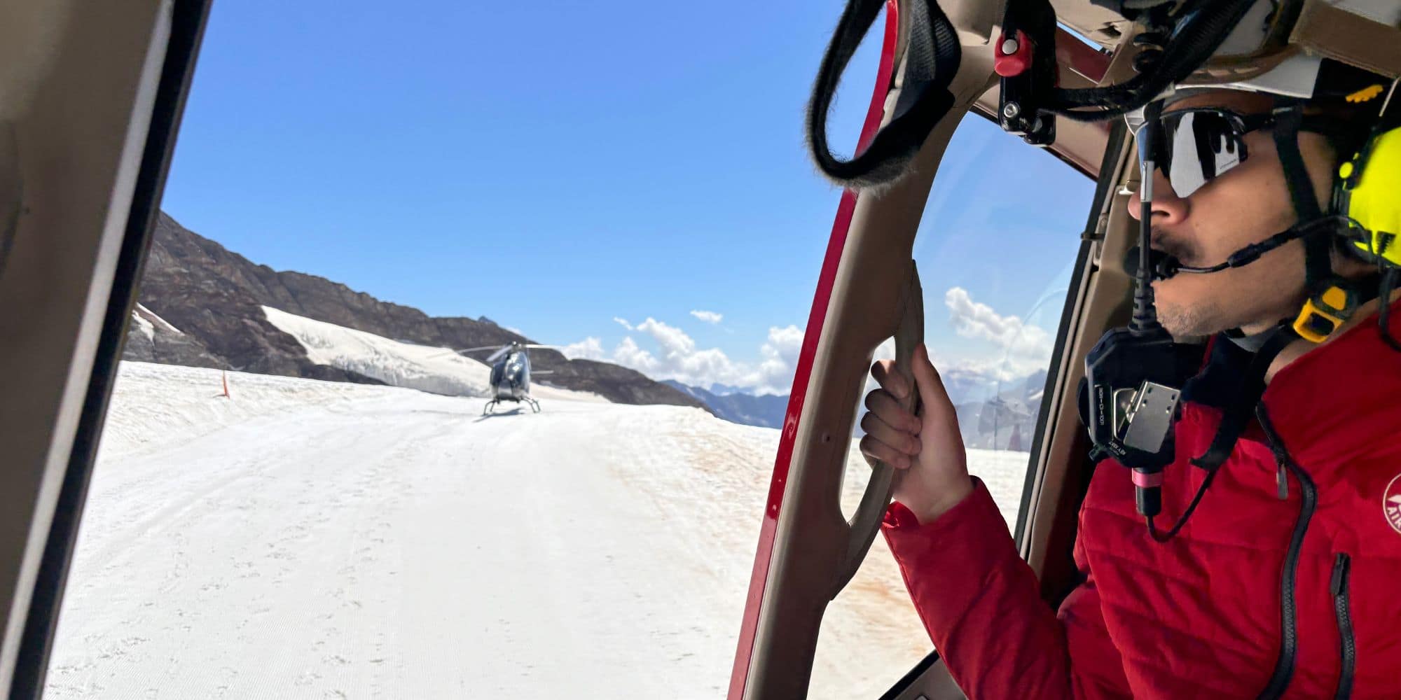 Pilot wearing a helmet and headset looks out from a helicopter cockpit toward another helicopter on a snowy mountain.