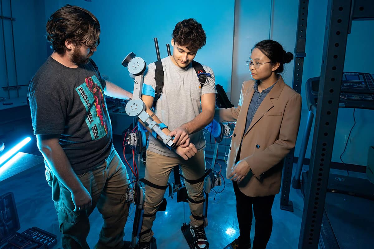 Student Edward Aleksi experiments with wearing a wearable robotic exoskeleton, designed to prevent common worker injuries. Alongside him are student Bryan Gonzalez (left) and Dr. Shuzhen Luo. (Photo: Embry‑Riddle/William Fredette-Huffman) 