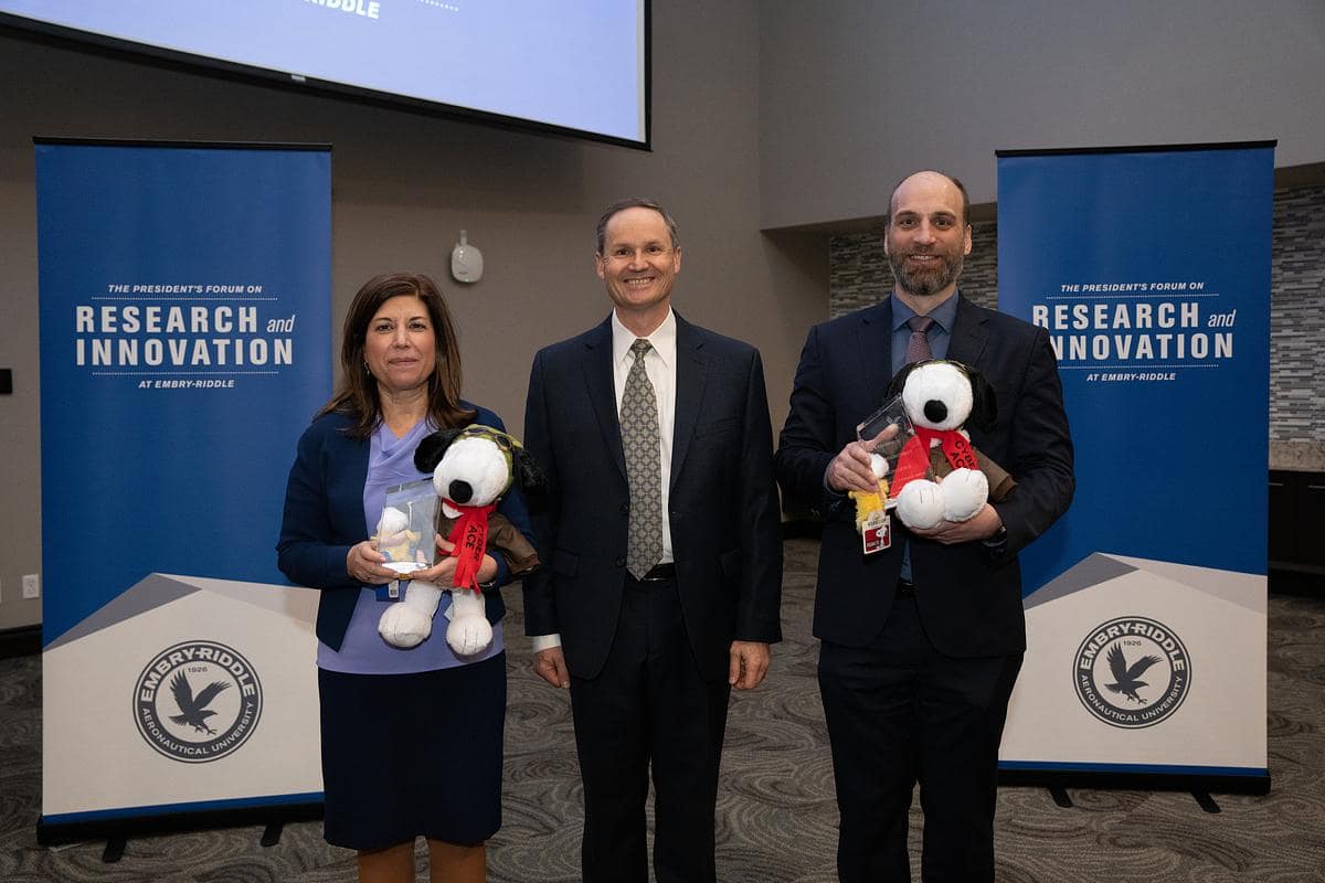 Three presenters standing between banners for the President’s Forum on Research and Innovation at Embry-Riddle, with two holding Snoopy-themed awards.