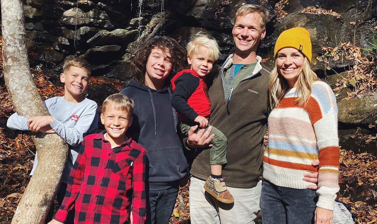 A family of six posing outdoors in front of a rocky waterfall with autumn leaves on the ground.