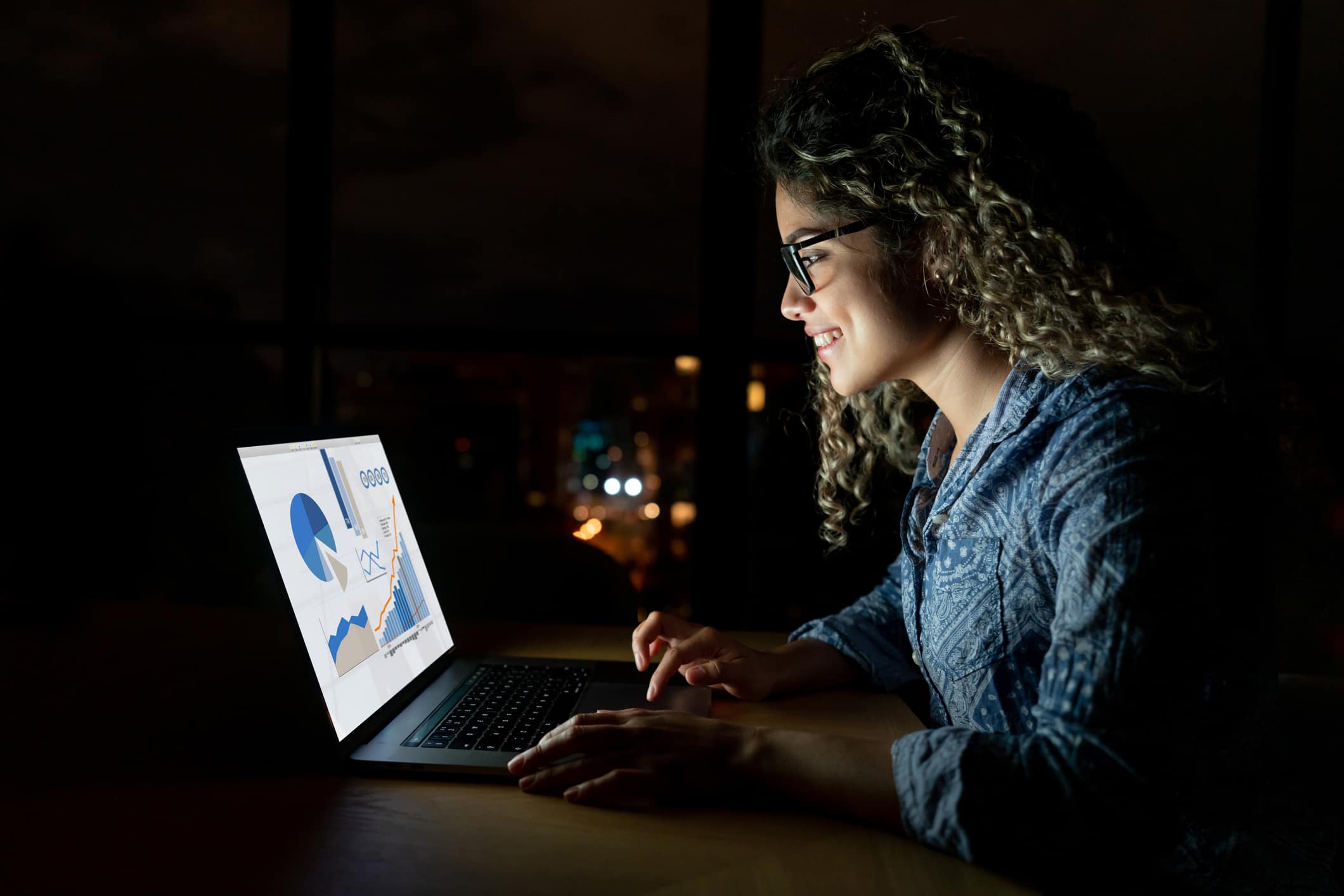 A student smiles broadly, using a laptop with data charts and graphs displayed on the screen, illuminated by the device in a dim room.